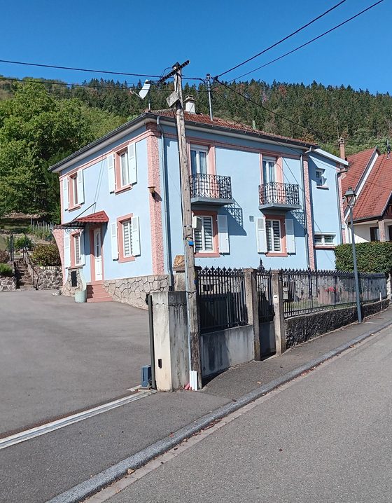 Façade de la maison à deux étages, enduit clair et volets, adossée à une colline boisée.