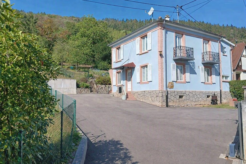 Façade bleue de la maison à deux étages, avec ses volets blancs et ses balcons en fer forgé, adossée à la colline boisée du Hahnenberg.